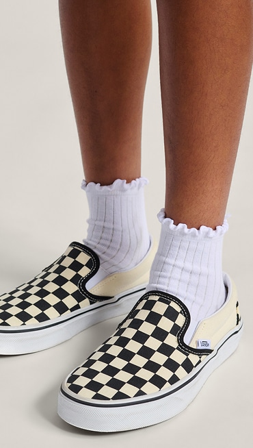 A knee-down shot of a kid wearing black and white Checkerboard Slip-On with white frilly socks in a photography studio.