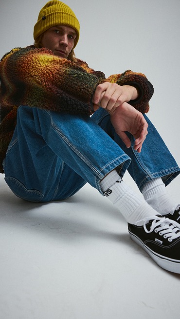 A young man in a sherpa jacket sits on the floor of a photography studio in blue jeans, white crew socks, and black Authentic.