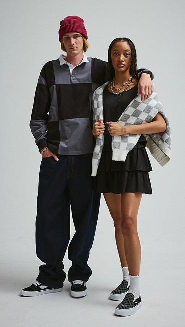 A young woman in a Checkerboard cardigan and a young man in a Checkerboard rugby polo pose together in a photography studio.