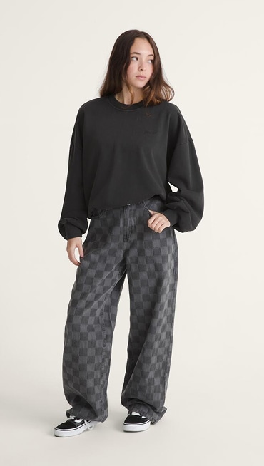 A young woman poses in a photography studio wearing black and gray Checkerboard pants, a black Script Logo Sweatshirt, and black and white Old Skool.