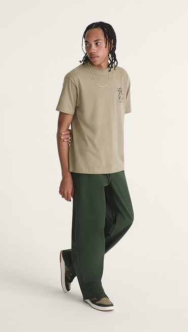 A young man poses in a photography studio wearing dark green Authentic Chino Loose pants and a brown shirt.