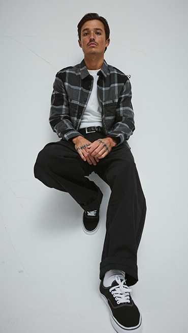 A man sitting in a photography studio wearing a gray Larkspur Plaid Flannel Shirt and black and white Authentic.