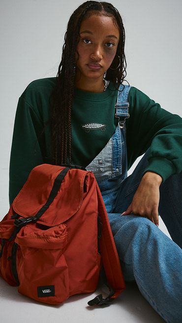 A young woman in a green Salton Pearls Crew Sweatshirt sits next to an orange backpack.