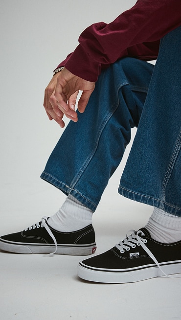 Close-up of someone wearing black Authentic with blue jeans and white Vans crew socks.