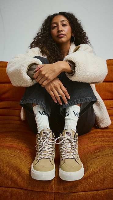 A young woman sits on an orange sofa wearing tan Sk8-Hi Insulated shoes, white Misty High Pile and black Puddle Pants.