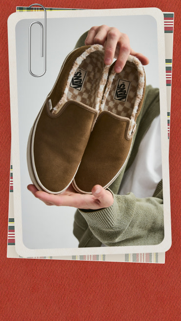 A young man holds up a pair of brown Classic Slip-On with fuzzy Checkerboard lining.