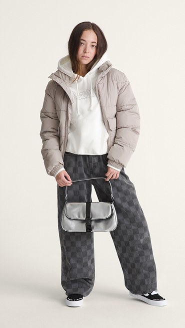 A young woman poses in a photography studio wearing black and white Sk8-Hi, a gray Misty High Pile, and black and gray Checkerboard Puddle Pants.