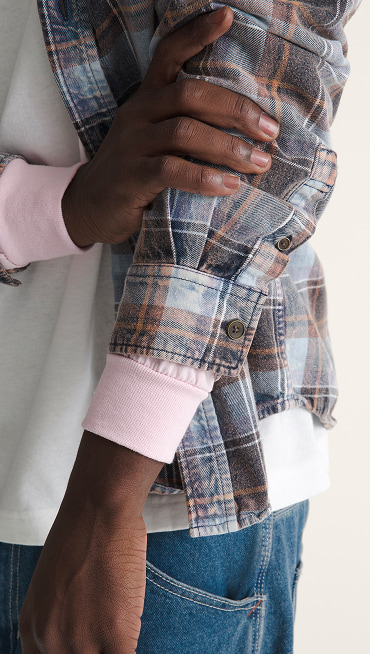 Close-up of a young man in a flannel holding his arm with a white tee and blue jeans.