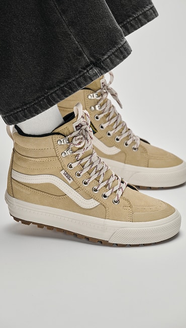 Close-up of a man sitting on a box wearing tan Sk8-Hi Insulated boots with black jeans in a photography studio.