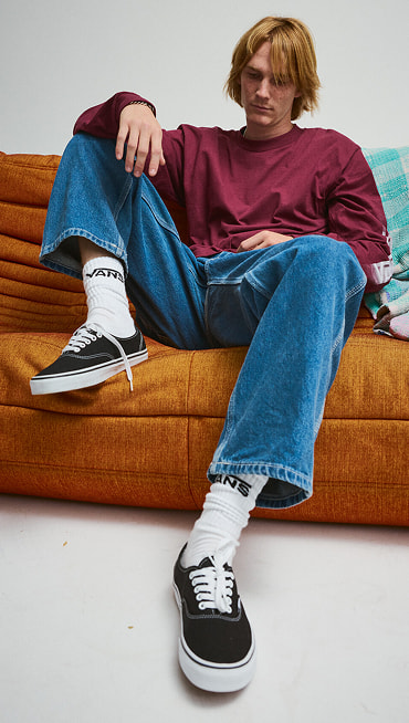 A young man poses on an orange sofa wearing black and white Authentic, blue jeans, and a red Blur Drop Long Sleeve T-Shirt.
