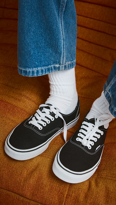 Someone wearing black and white Authentic on an orange sofa with blue jeans and white crew socks.
