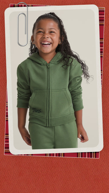 A young girl is smiling, wearing a green Little Kids Unicorn Zip Hoodie and matching sweatpants.