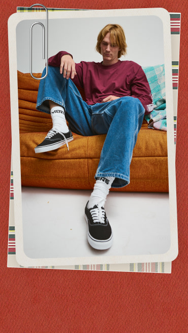 Man wearing blue jeans, Vans white and black socks, black and white Vans Authentic Shoes and a red shirt on while sitting on a couch.