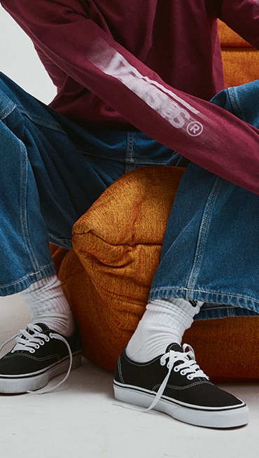 A waist-down shot of a young man sitting on an orange sofa wearing black and white Authentic shoes with blue jeans and white Vans crew socks.