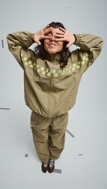 A young woman puts her hands over her face wearing the yellow and brown Ronnie Track Jacket and Pants in a photography studio.