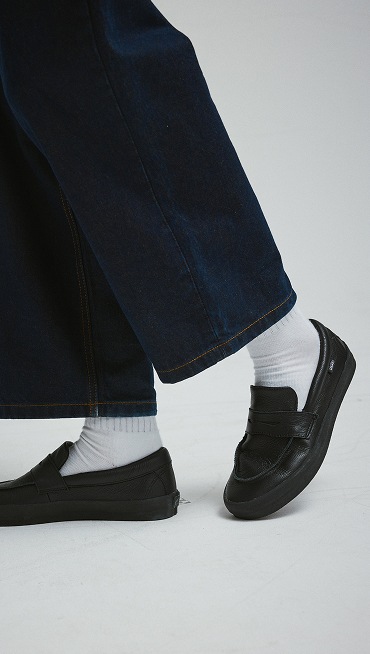 A knee-down shot of a young man wearing black Skate Loafers with white socks and dark blue jeans.