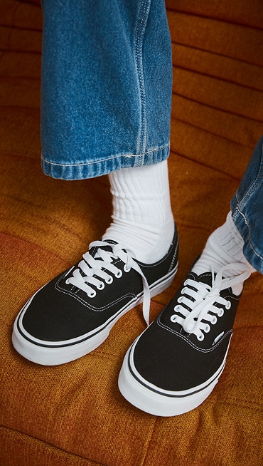A knee-down shot of a young man sitting on an orange sofa wearing black and white Authentic shoes with blue jeans and white Vans crew socks.