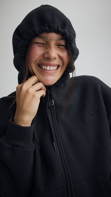 Medium shot of a woman smiling in a Salton Full Zip Fleece in Black in a grey photography studio.