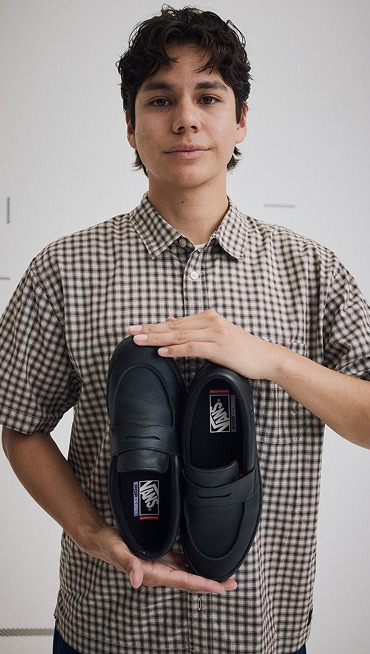 Medium shot of Nick Michel holding a pair of Skate Loafers in Black while wearing a Monte Plaid Button Up Shirt in Egret White.