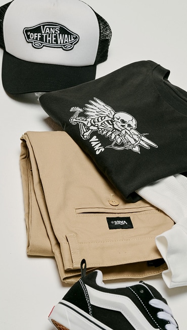 A full outfit is laying on the floor.. Complete with a pair of Knu Skool shoes, Kids Authentic Chino Loose Pant, a black t-shirt and a black and white hat.