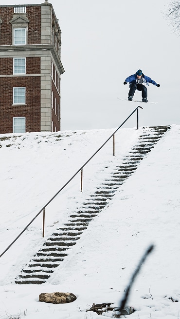 Image of Vans Snowboarder Jill Perkins in the air as she's about to land on a rail that goes all the way down a flight of snowy stairs.