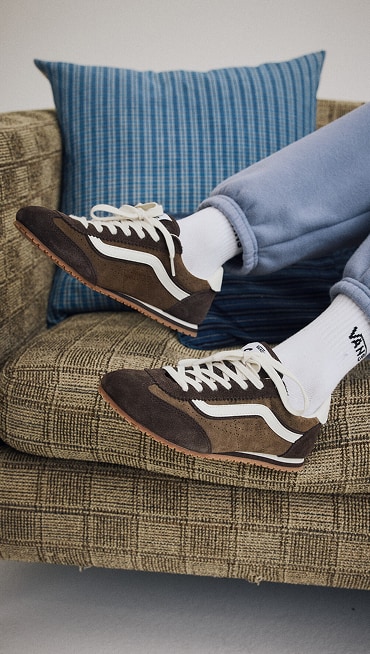 A knee-down shot of someone wearing demitasse brown Super Lowpro, light blue sweats, and white Vans crew socks on a retro-looking sofa.