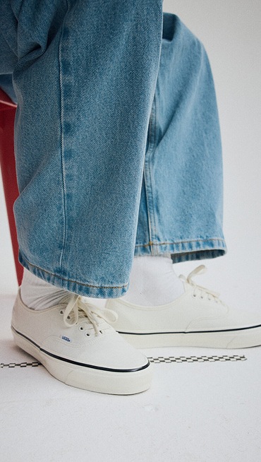 Close up of someone wearing the Premium Authentic Shoe in Marshmallow White while sitting on a red chair in a grey photography studio.