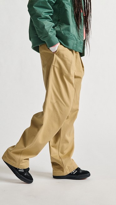 A person walking on a white studio backdrop wears a green jacket, tan pleated pants, and Upland sneakers in black.