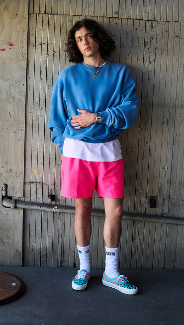 An image of a person standing in front of a wooden wall. They are wearing a blue crewneck sweater, a white shirt underneath, pink shorts, mid-calf white socks, and Premium Authentic Shoes in Cheetah Pool Blue Multi.