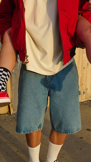 A man wearing baggy denim shorts past the knee carrying a friend wearing red Authentic shoes and Checkerboard socks.