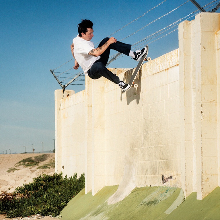 Vans Skateboarder Elijah Berle is skating on the edge of a cement wall with barbed wire fencing. The background is blue sky and a sandy beach.
