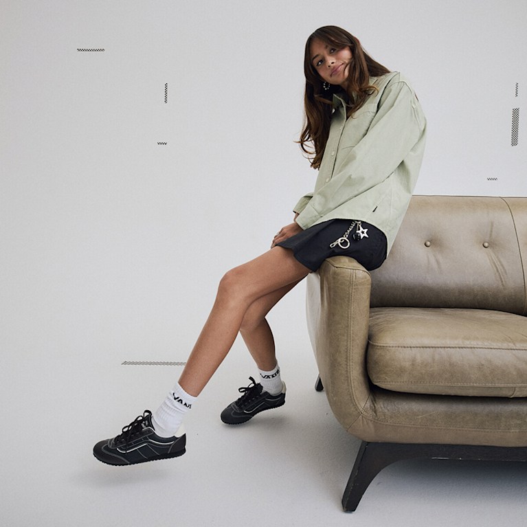 Vans Surf Team Rider Maddie Miller leans on the arm of a leather sofa wearing a black Authentic Chino Skirt and a green Utility Shirt.