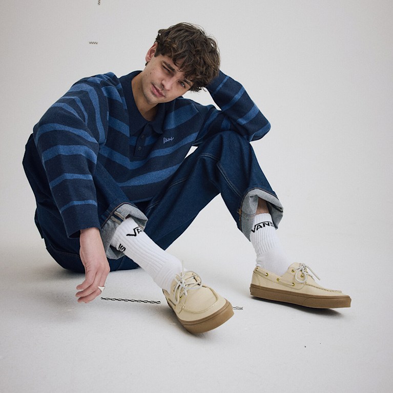 A young man sits on the floor of a photography studio wearing a blue Davis Stripe Polo Sweater and cream 2-Eyelet Boat shoes with white crew socks.
