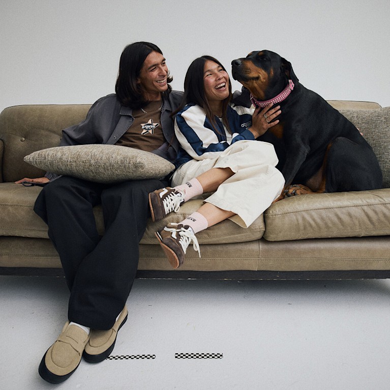 Wide shot of a man and a woman wearing Vans shoes, pants, shirts and jackets while sitting on a couch with a dog who is getting some pets and smiles.