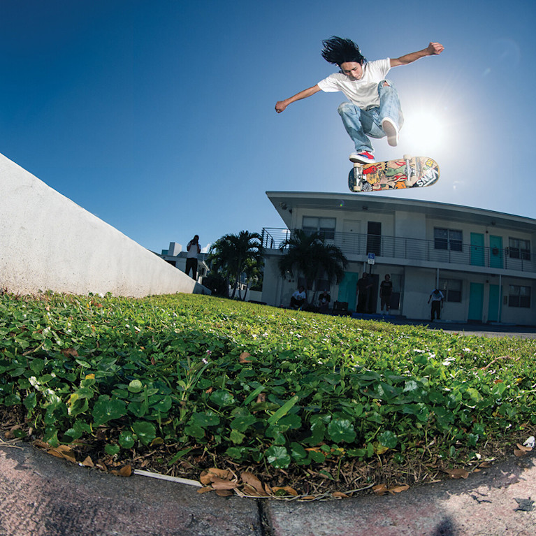 Panoramic shot of Daiki Hoshino outside skateboarding mid jump from a cement ramp over grass onto a driveway wearing the Blue / Red Skate Era Shoe.