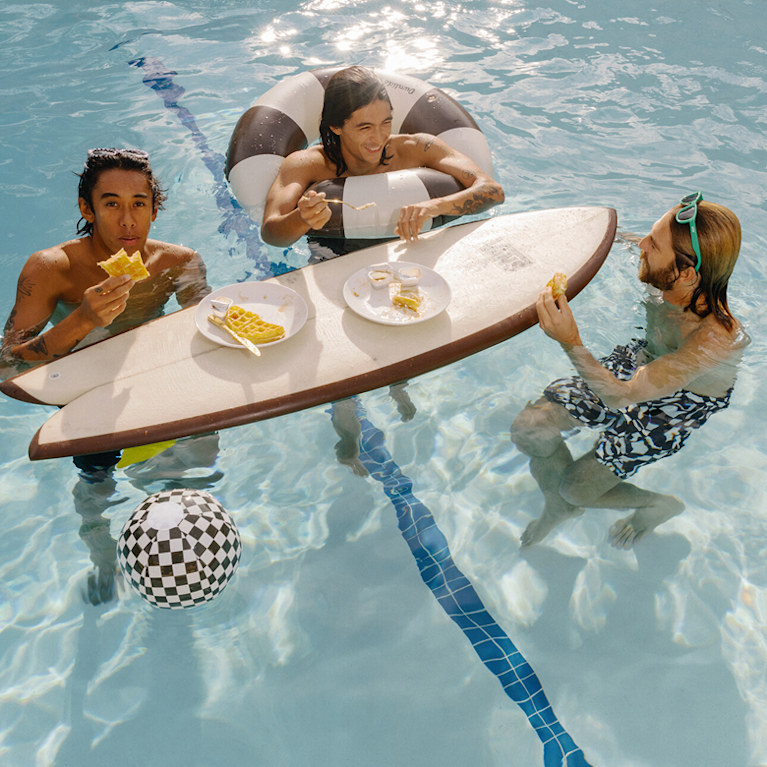 Trois jeunes hommes dans une piscine portent des boardshorts Vans et mangent des gaufres posées sur une planche de surf Vans.