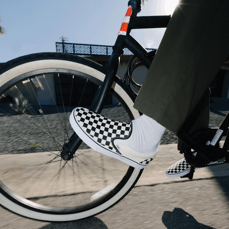 Close-up of someone biking outside wearing white and black Checkerboard Classic Slip-On shoes.