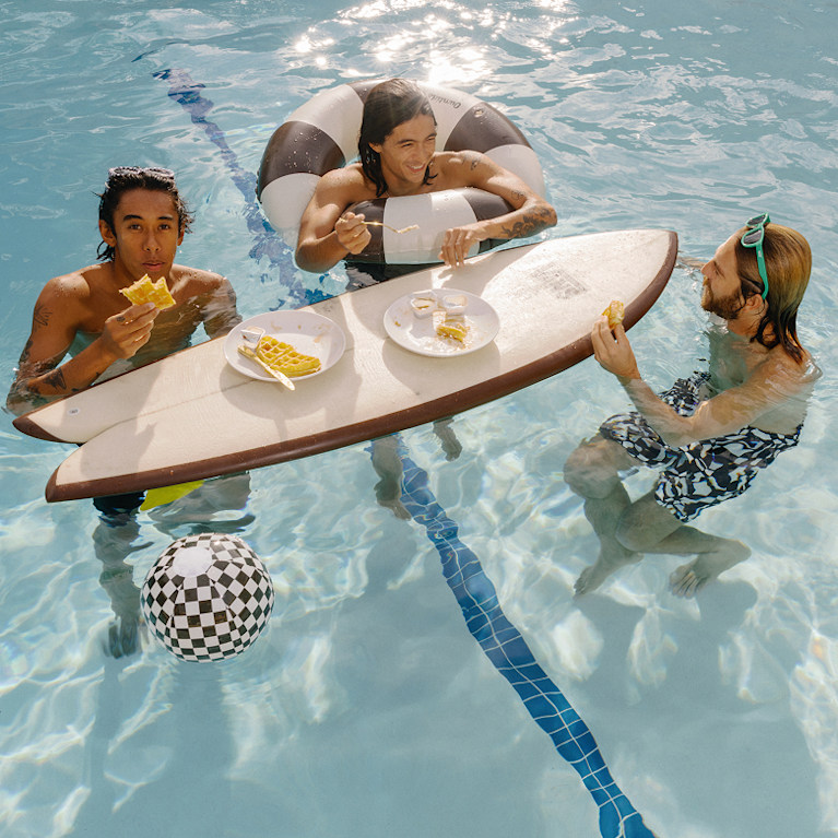 A group of young men stand in a pool eating waffles off a Vans surfboard wearing Vans boardshorts and sunglasses.