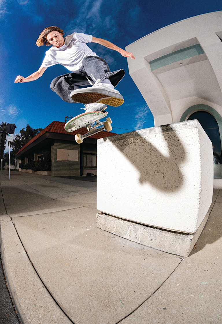 A well-lit shot of Curren jumping to grind on a concrete block wearing a white t-shirt, black jeans, and white Curren shoes.