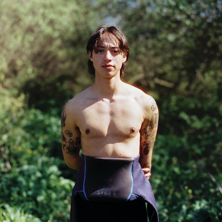 A surfer with their wetsuit down to their ribs stares into the camera