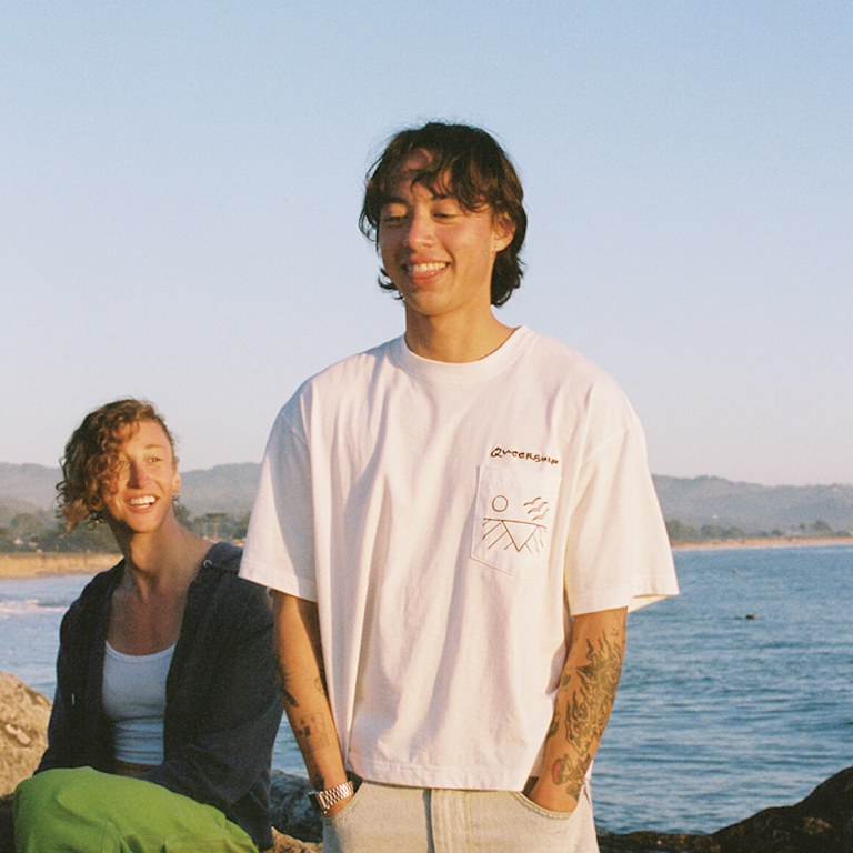 Two people are shown on the beach at sunset. One person is standing wearing a white t-shirt with the phrase "Queer Surf" over the pocket.