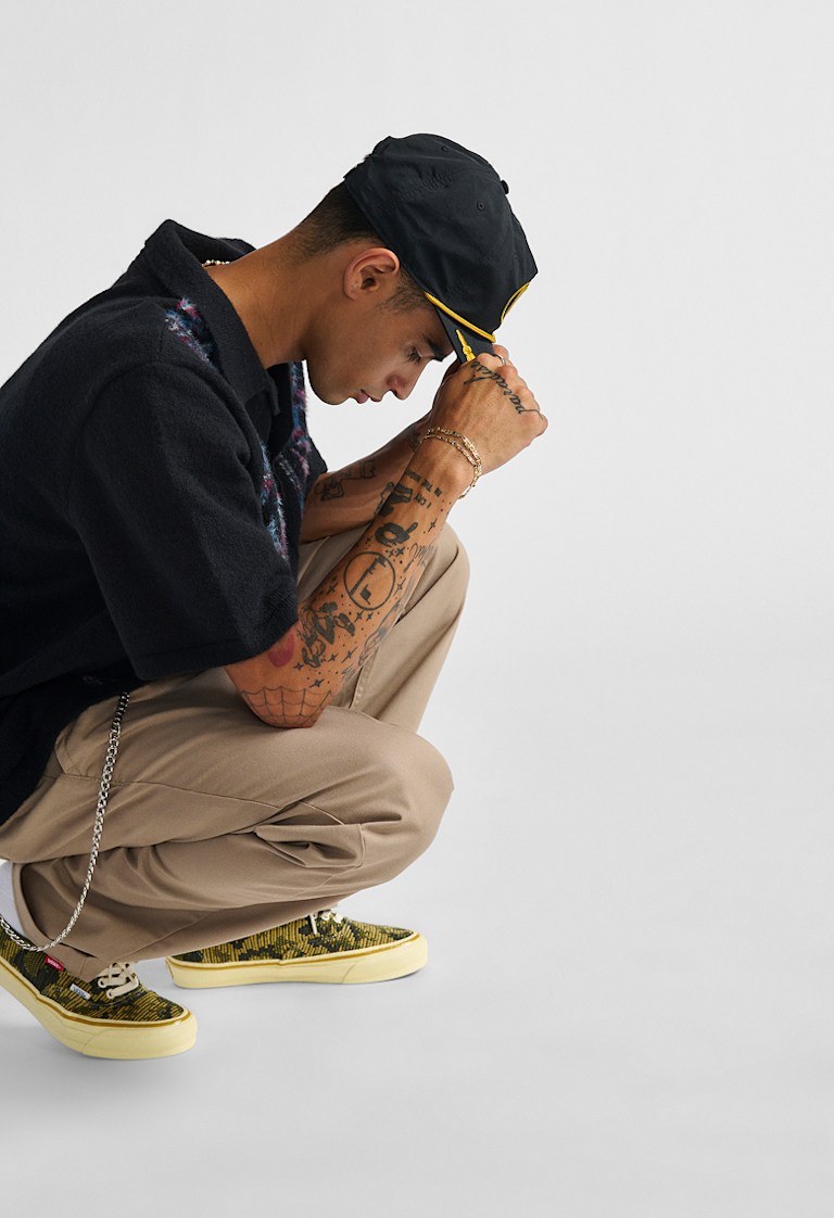 Studio shot of a man posing with his Scrambled Vans Snapback Hat, styled with a black shirt, khakis, and Vans Premium Authentic shoes.