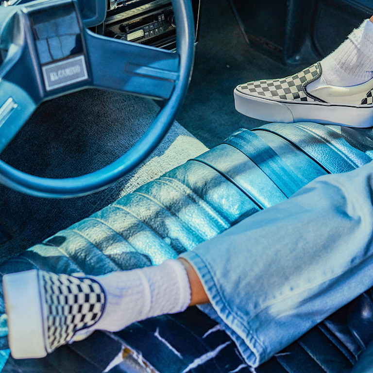 A waist-down shot of someone in a retro car wearing blue jeans and black and white Checkerboard Classic Slip-On.