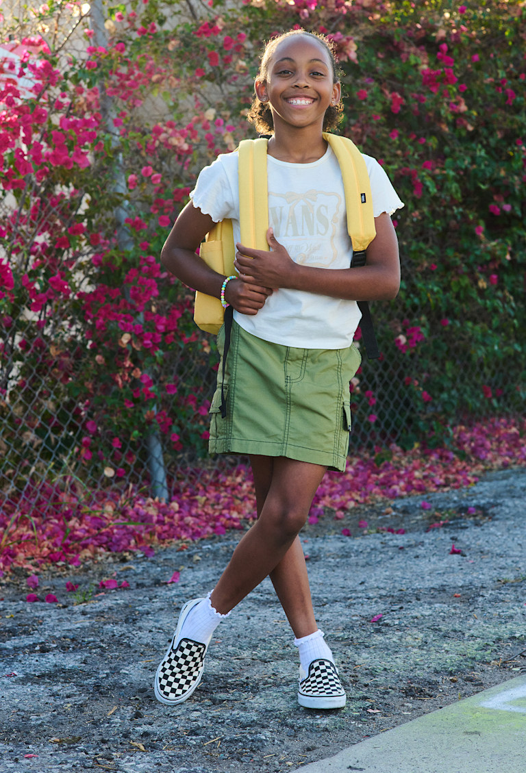 A young girl poses with her banana cream yellow Kids Old Skool Grom Backpack and Loden Green Kids Sidewalk Cargo Skirtin front of a rose bush.