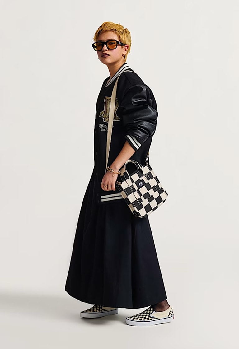 A woman poses in a photography studio wearing a black varsity-style jacket, black skirt, and a black and white Lil Pergs Tote Bag.