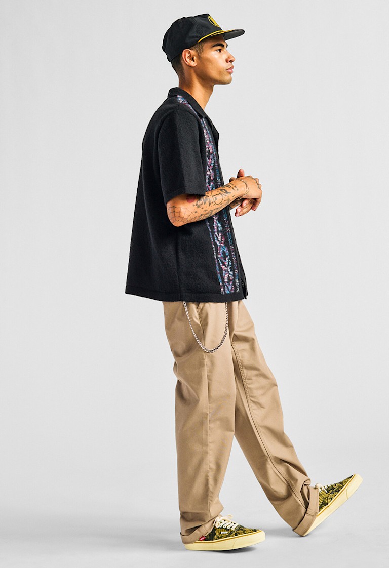 A man steps and poses in a photography studio wearing a black Wheeler Sweater Shirt, brown chinos, and Antique Gold and Green Premium Authentic.