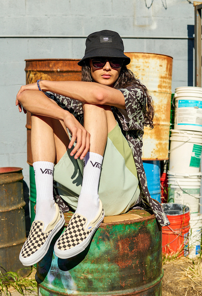 A young man sits on a rusty oil drum wearing black and white Checkerboard Classic Slip-On with white Vans crew socks and a black Vans bucket hat.