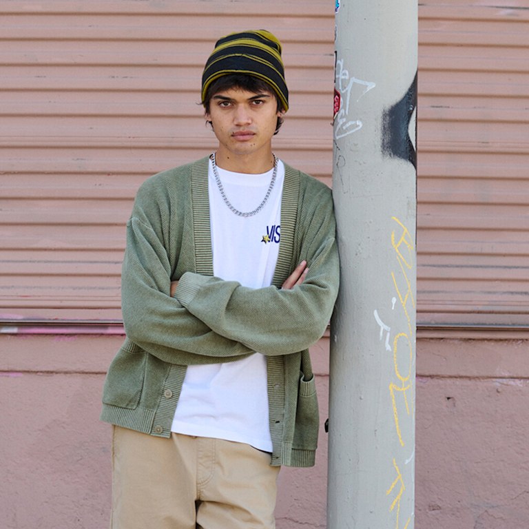 A young man scowls at the camera wearing a white Vans t-shirt and a moss green cardigan as he leans against a metal pole.