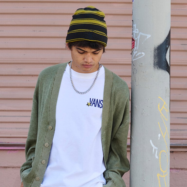 A young man looks at his feet wearing a white Vans t-shirt and a moss green cardigan as he leans against a metal pole.