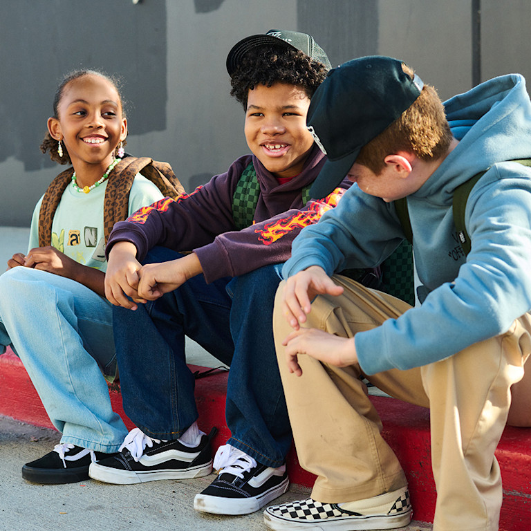 Three young kids sit on a curb wearing Vans clothes, backpacks, Old Skool, Knu Skool, and Checkerboard Classic Slip-On.
